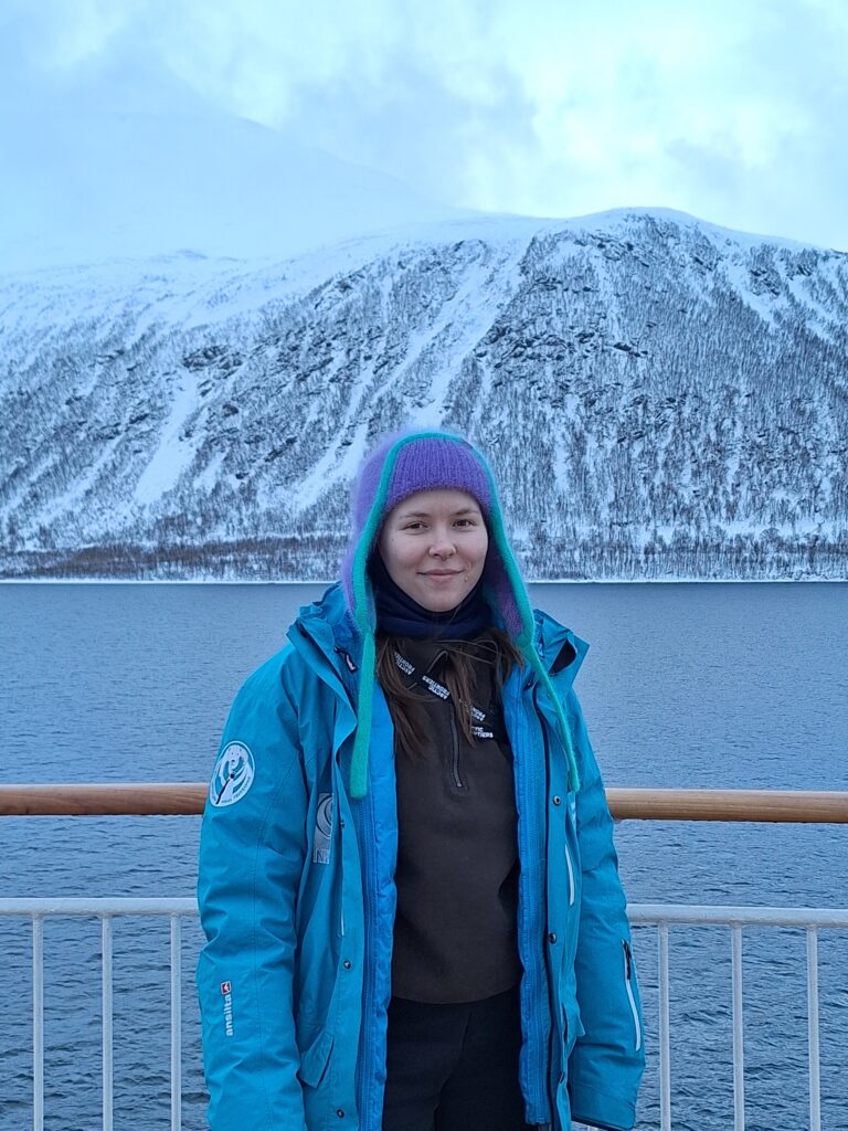 Olga posing in front of a reeling on board of a vessel. Snow-powdered mountains in the background.