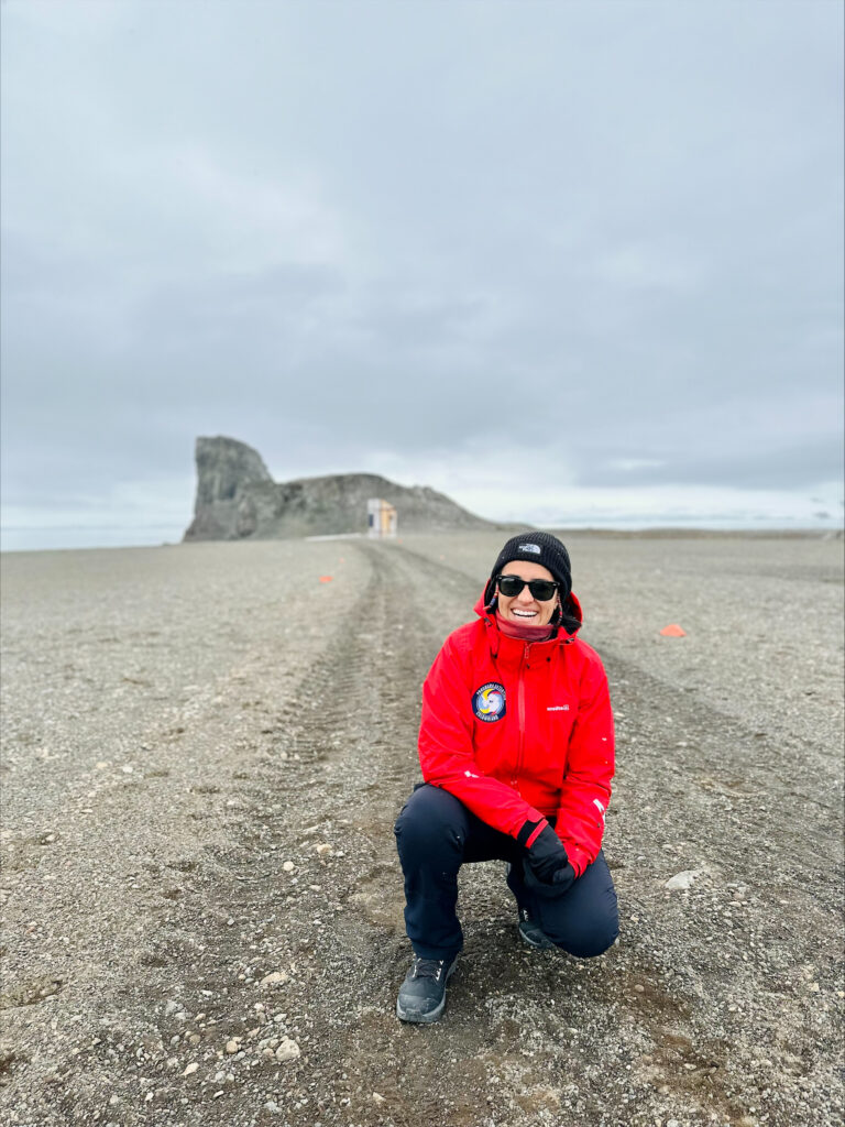 Natalia, in red polar parka, kneels in front of a rock formation on a gravelly beach.