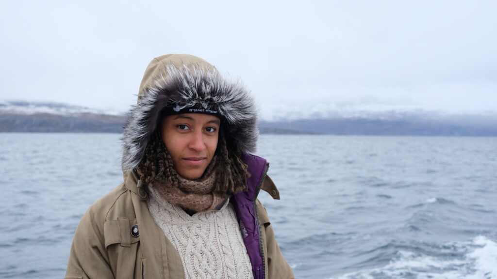 Myrah, in a hooded parka, is standing in front of a body of water, with snow-capped hills in the background. Photo was taken outside Nain, Nunatsiavut (Canada). Myrah was on Willie Fox's boat for a benthic survey in 2022.